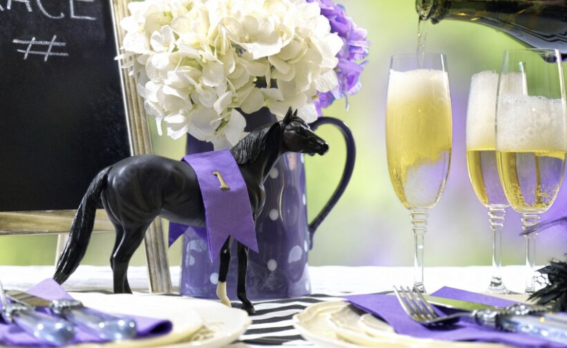 Derby table setting with a model racehorse, purple ribbon, flowers and sparkling drinks